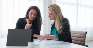 Two women, dark hair and blonde long hair - discussing onboarding at a desk. Desk and background are a fresh white with filtered blinds behind the women.
