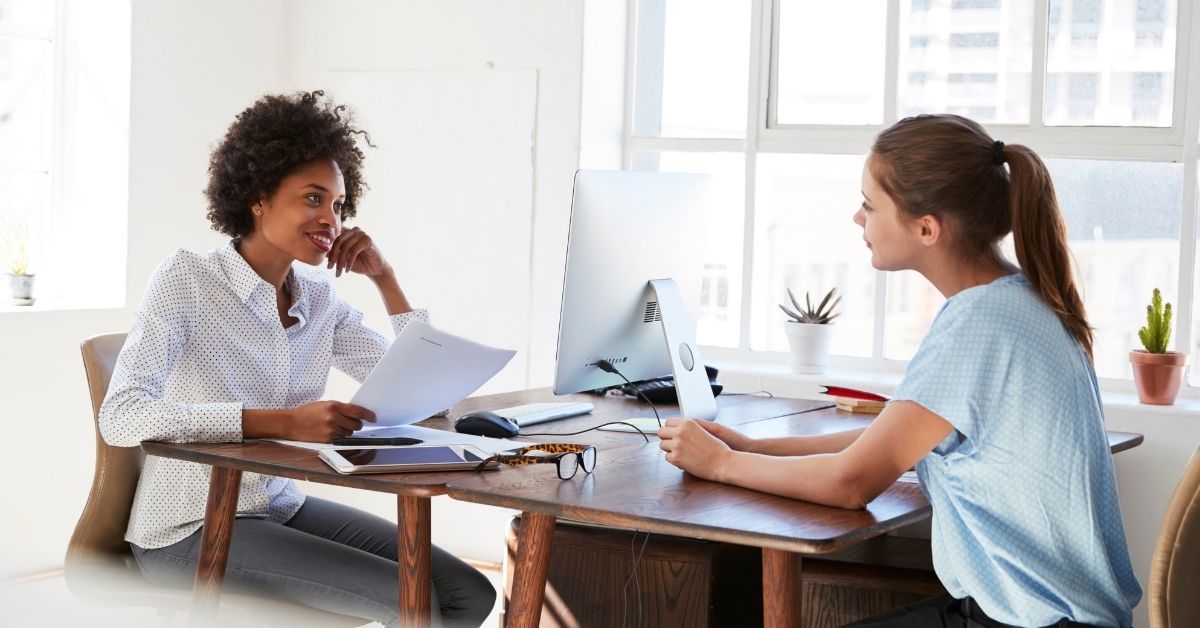 Two women speaking across a desk. The black woman is holding a piece of paper, the white woman listening intently, office is a light fresh background