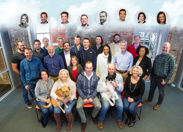 A team gathered for a photo in an office setting, with grey floor and brick walls. Behind the team are two windows and above the team are clouds with people's faces in them.