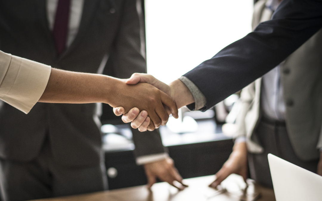 Business setting with a man and woman shaking hands over a desk with male and female witnesses. No faces are showing.